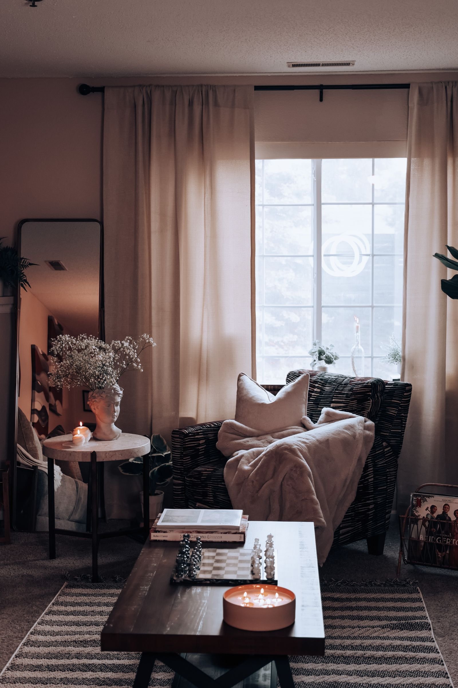 A warm living room in the evening with candles, a soft throw over an armchair, a floor mirror and layered textiles